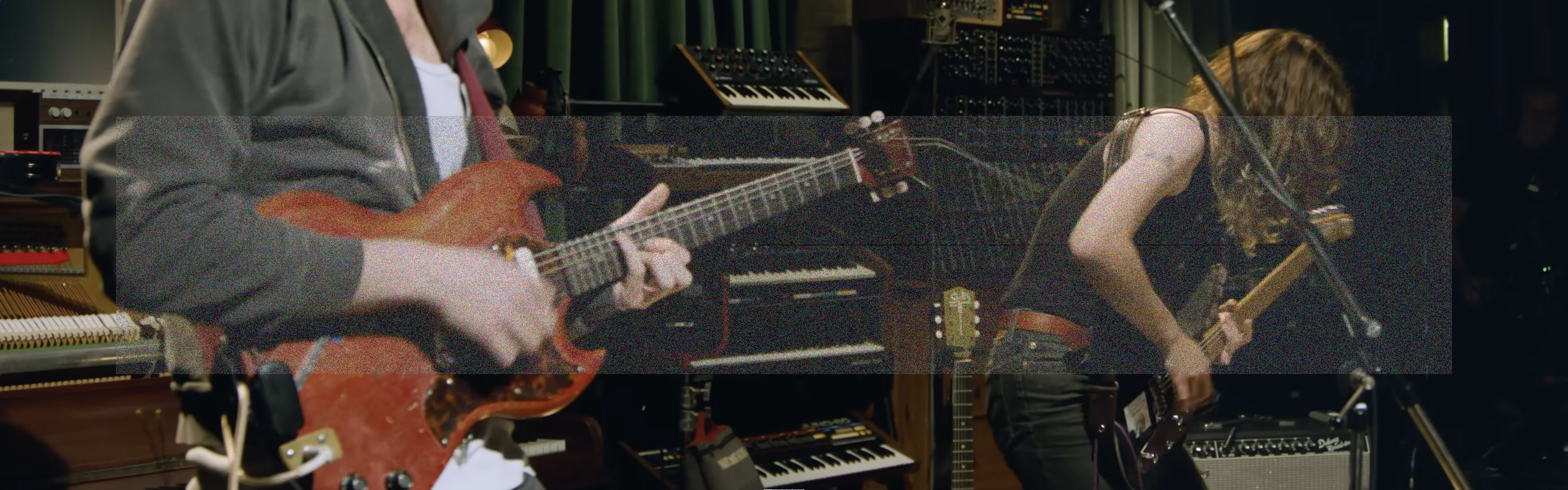 An ultrawide landscape photo cropping Cameron Winter and Emily Green playing electric guitars with studio instruments in the background. A wide rectangle of fine grainy static lightly veils the center of the image.