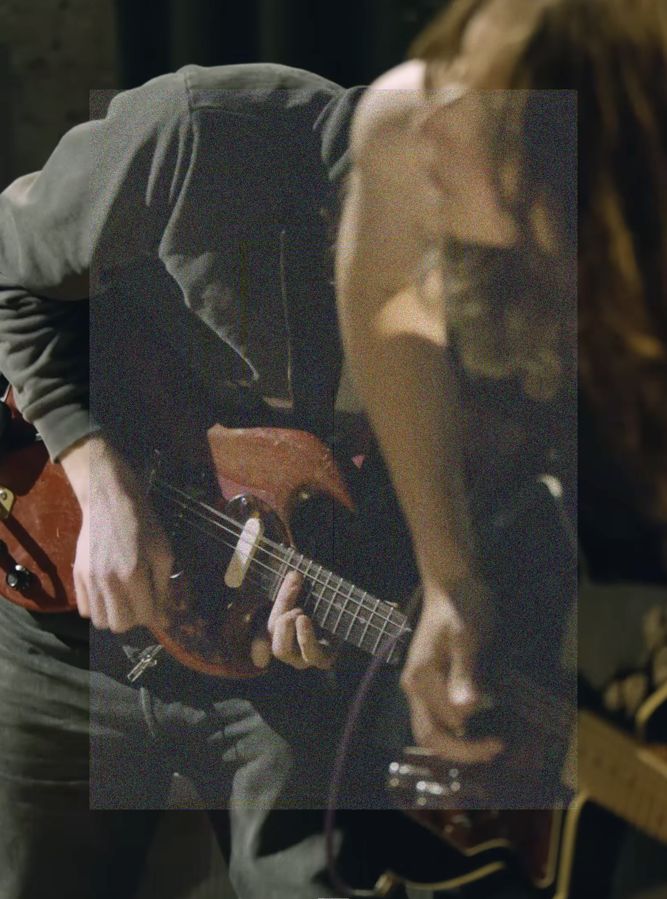 A cropped vertical shot of Cameron Winter with Emily Green out of focus in the foreground, both leaning downward in the same direction playing their guitars intensely. The shot framing lines them up at the same downward angle, with both bodies and guitars blending into each other. A tall rectangle of fine grainy static lightly veils the center of the image.