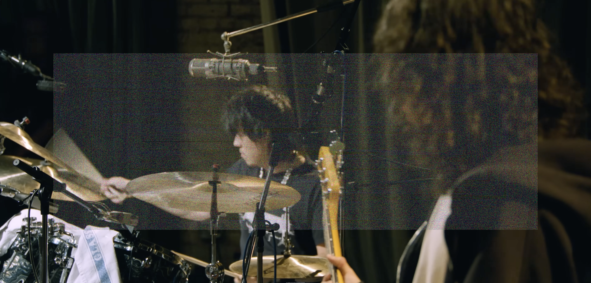 A wide shot of Max Bassin playing drums, stick actively hitting a ride cymbal, with Dominic DiGesu out of focus and facing away from camera in the foreground with the neck of his bass in view. A wide rectangle of fine grainy static lightly veils the center of the image.