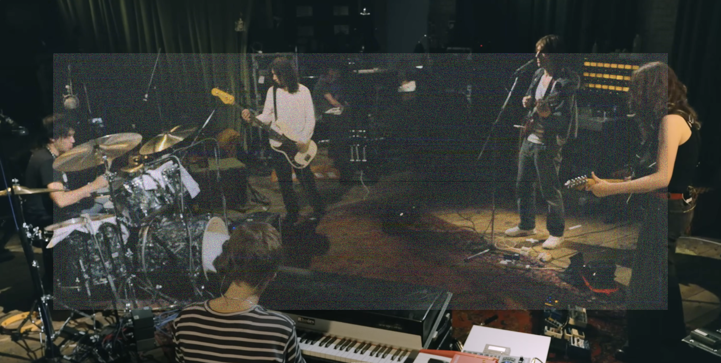 A high angle wide shot of the Geese band playing in the studio. Clockwise, Max Bassin on drums, Dominic DiGesu playing bass, Cameron Winter on guitar and singing, Emily Green playing guitar, and the back of Sam Revaz playing a Fender Rhodes. A wide rectangle of fine grainy static lightly veils the center of the image.