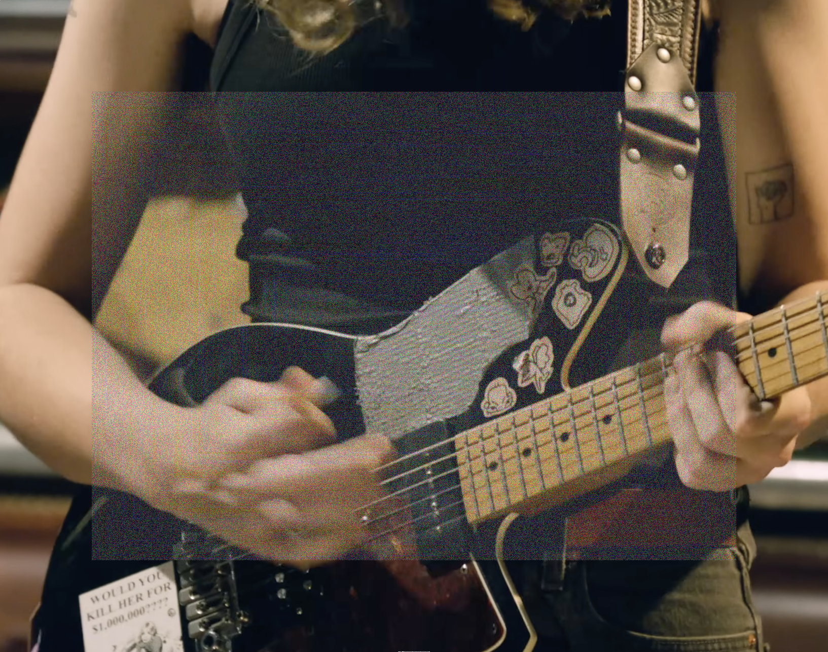 A rectangular image, close up crop of Emily Green's hands playing guitar. On here guitar is a sticker that says 'Would you kill her for $1,000,000????'. A landscape rectangle of fine grainy static lightly veils the center of the image.