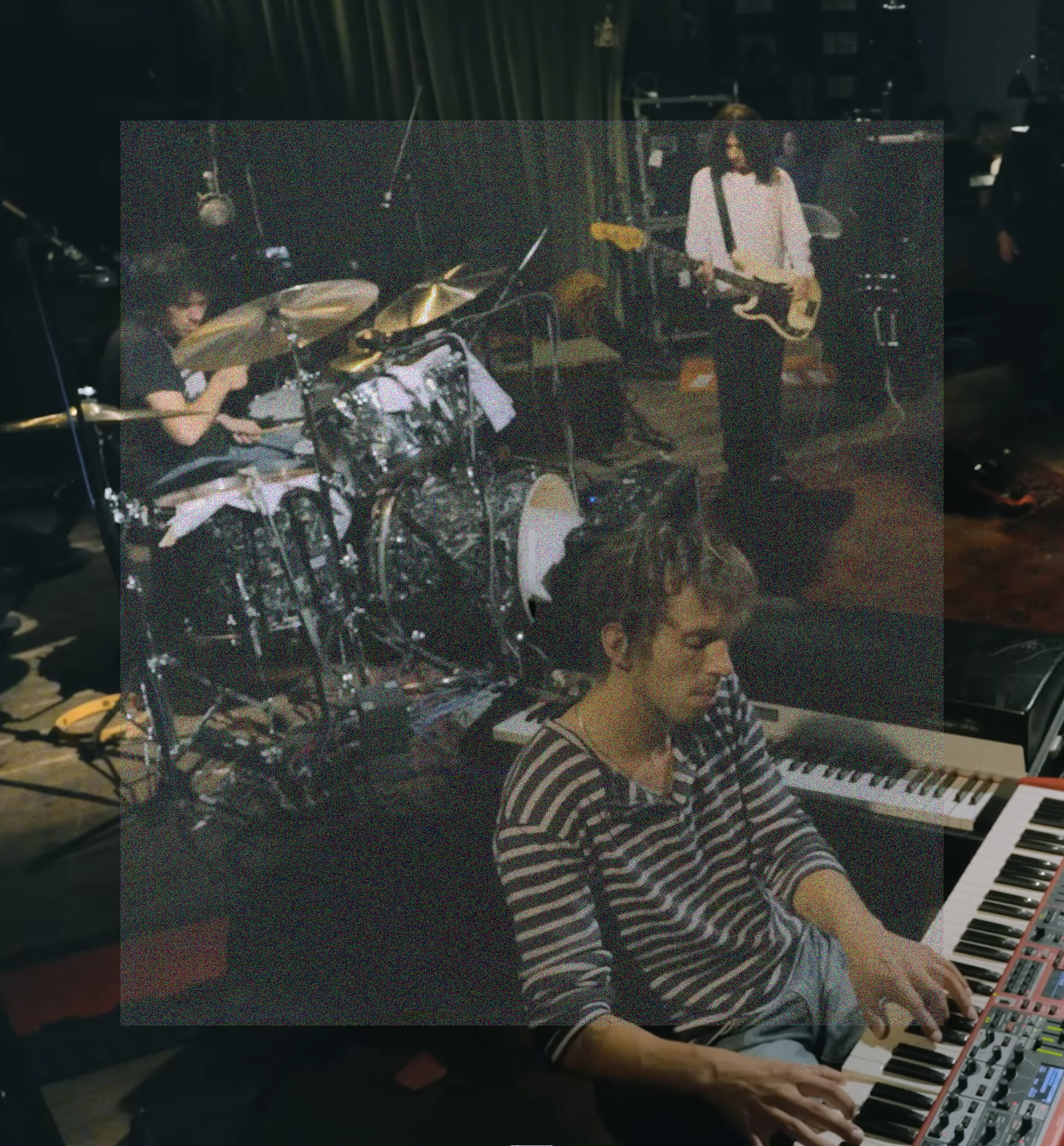 A vertical rectangular image, high angle shot of of Sam Revaz playing an electronic piano with Max Bassin and Dominic DiGesu playing drums and bass in the background. A rectangle of fine grainy static lightly veils the center of the image.