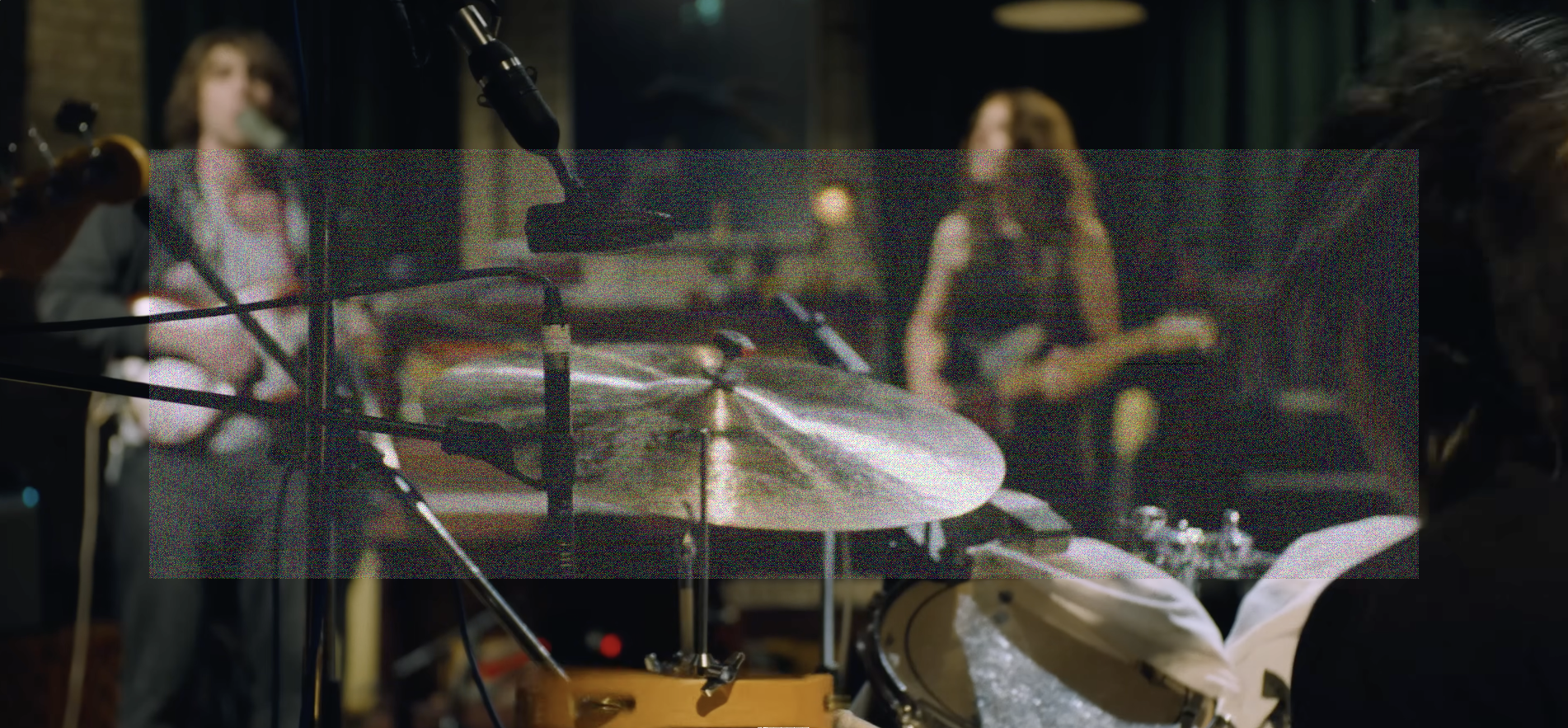 A wide angle over the shoulder shot behind Max Bassin and his drum kit. In the background and out of focus is a singing Cameron Winter and Emily Green playing guitar. A rectangle of fine grainy static lightly veils the center of the image.