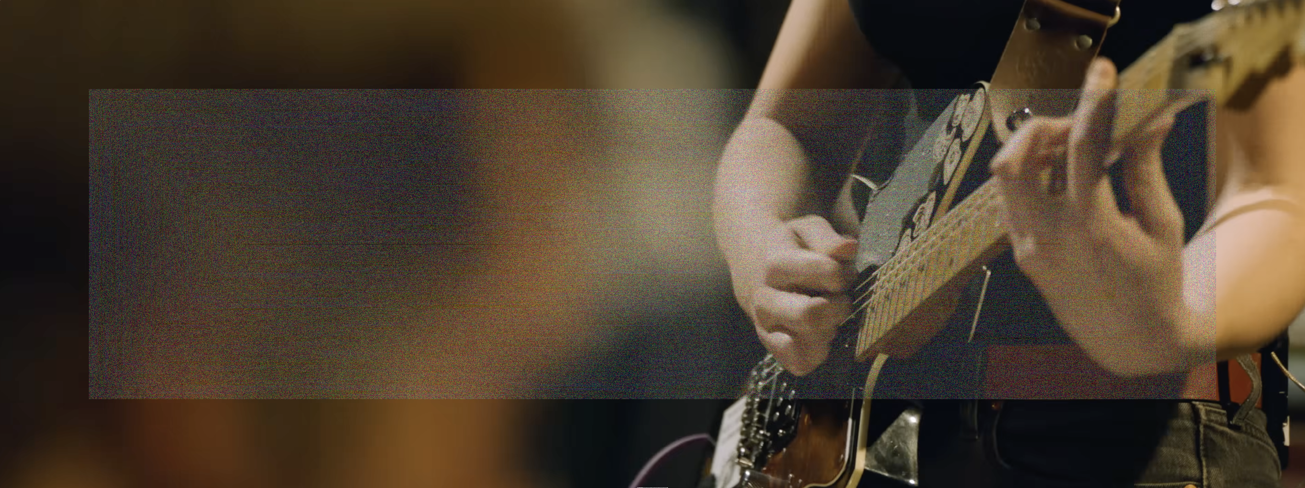An over the shoulder wide angle shot of Emily Green's hands playing a power chord on her guitar on the right, and the back of Sam Revaz's head out of focus in the foreground on the left. A wide rectangle of fine grainy static lightly veils the center of the image.