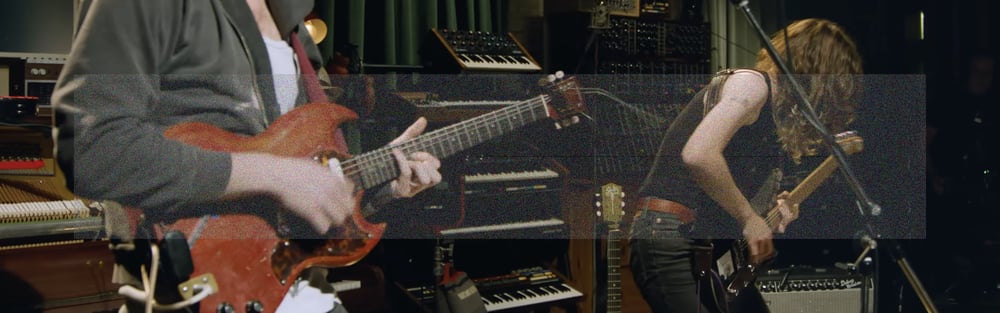 An ultrawide landscape photo cropping Cameron Winter and Emily Green playing electric guitars with studio instruments in the background. A wide rectangle of fine grainy static lightly veils the center of the image.