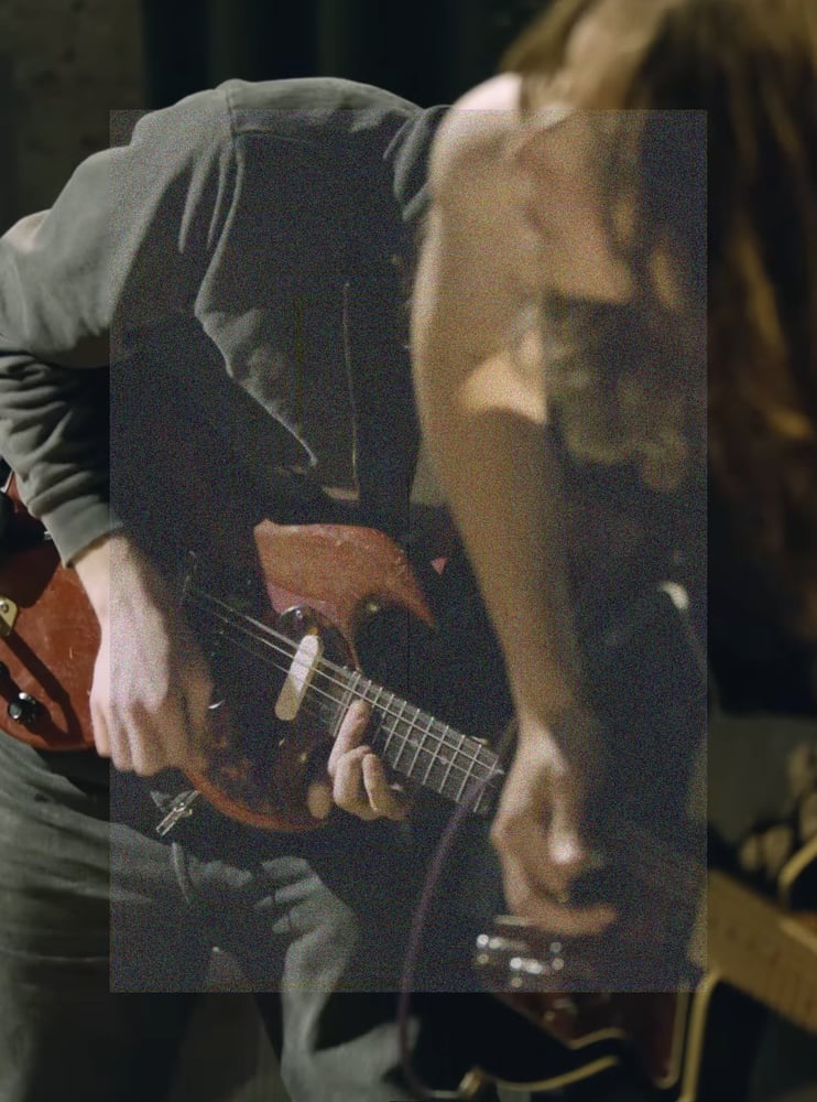 A cropped vertical shot of Cameron Winter with Emily Green out of focus in the foreground, both leaning downward in the same direction playing their guitars intensely. The shot framing lines them up at the same downward angle, with both bodies and guitars blending into each other. A tall rectangle of fine grainy static lightly veils the center of the image.