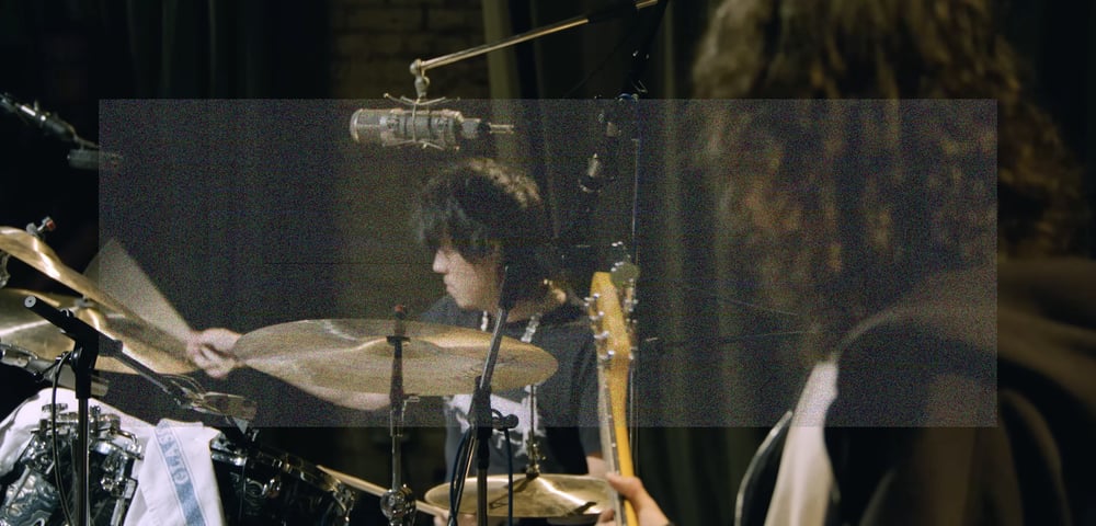A wide shot of Max Bassin playing drums, stick actively hitting a ride cymbal, with Dominic DiGesu out of focus and facing away from camera in the foreground with the neck of his bass in view. A wide rectangle of fine grainy static lightly veils the center of the image.