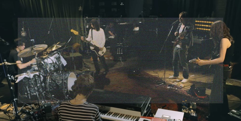 A high angle wide shot of the Geese band playing in the studio. Clockwise, Max Bassin on drums, Dominic DiGesu playing bass, Cameron Winter on guitar and singing, Emily Green playing guitar, and the back of Sam Revaz playing a Fender Rhodes. A wide rectangle of fine grainy static lightly veils the center of the image.