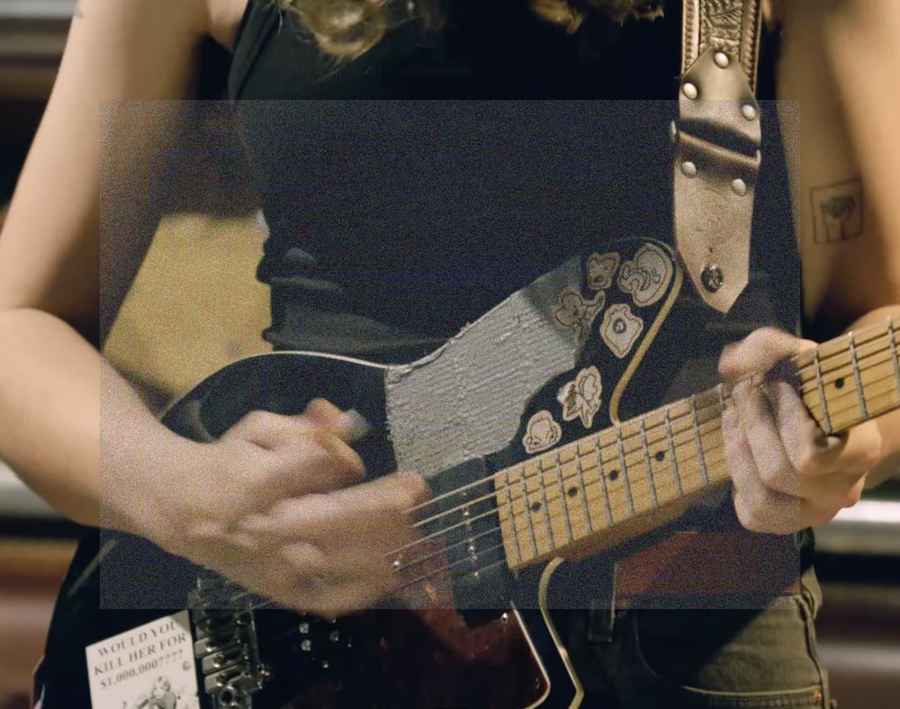 A rectangular image, close up crop of Emily Green's hands playing guitar. On here guitar is a sticker that says 'Would you kill her for $1,000,000????'. A landscape rectangle of fine grainy static lightly veils the center of the image.