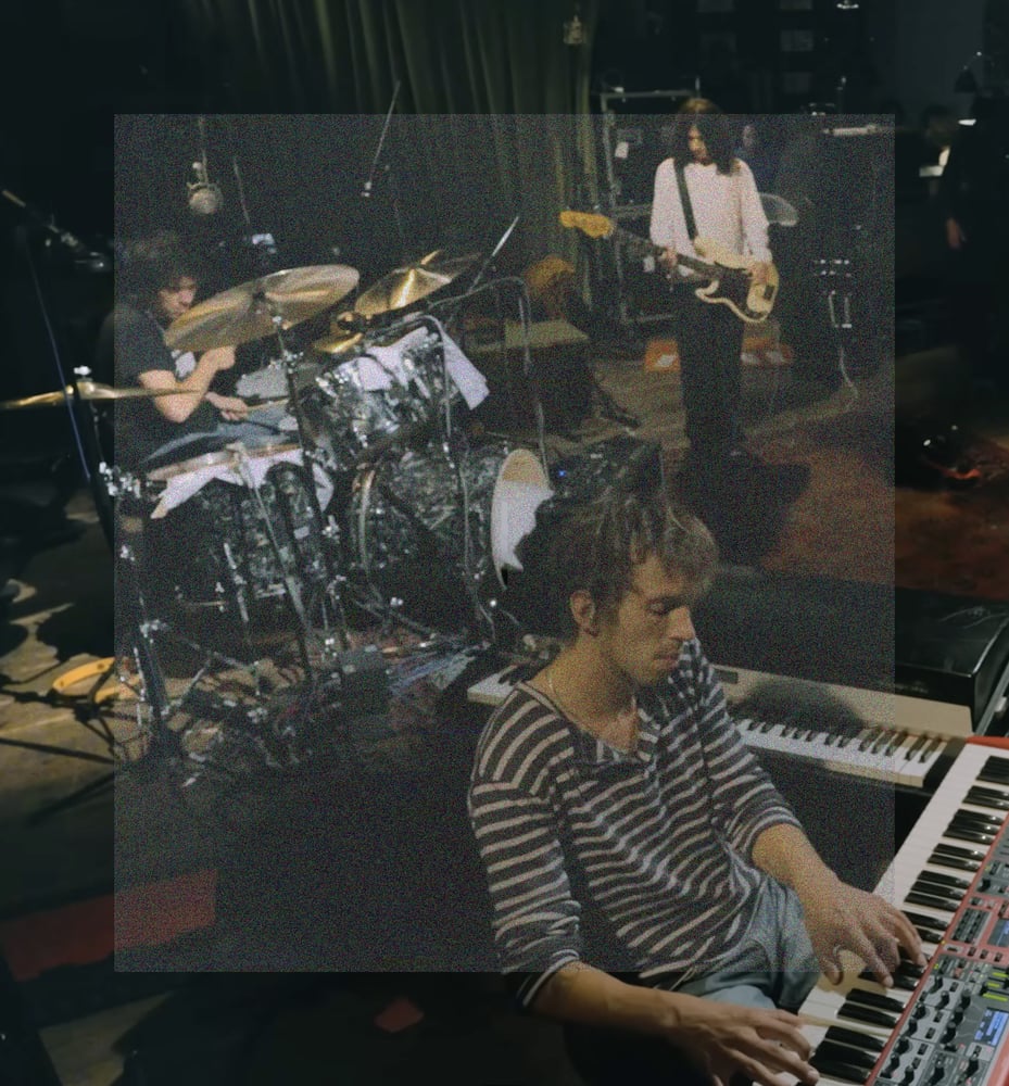 A vertical rectangular image, high angle shot of of Sam Revaz playing an electronic piano with Max Bassin and Dominic DiGesu playing drums and bass in the background. A rectangle of fine grainy static lightly veils the center of the image.