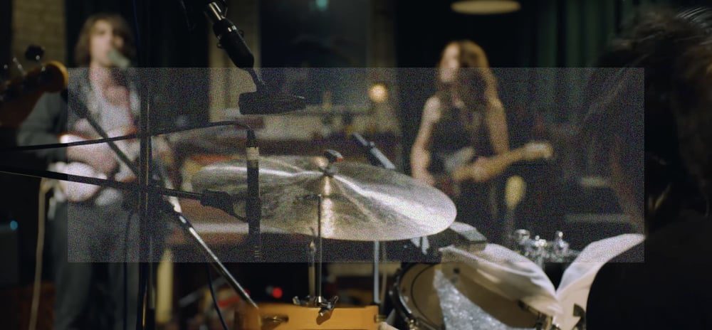 A wide angle over the shoulder shot behind Max Bassin and his drum kit. In the background and out of focus is a singing Cameron Winter and Emily Green playing guitar. A rectangle of fine grainy static lightly veils the center of the image.