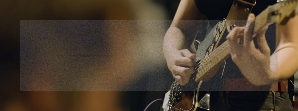An over the shoulder wide angle shot of Emily Green's hands playing a power chord on her guitar on the right, and the back of Sam Revaz's head out of focus in the foreground on the left. A wide rectangle of fine grainy static lightly veils the center of the image.