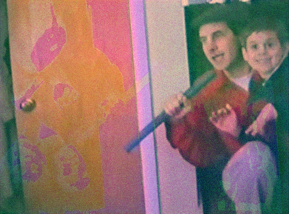 A boy leans out from behind a bedroom door beside a man holding him, both smiling at the camera. The door panel on the left is posterized into flat bands of orange, pink and magenta, and dense colored grain sits over the frame.