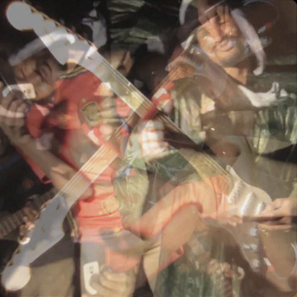 A warm square photograph of Mdou Moctar playing an electric guitar in formal Saharan clothing, surrounded by people dancing outdoors in the dark under harsh directional lighting. A translucent ghost of the same scene is laid over itself, blending the warm oranges, browns, and earth tones into a diffuse dreamlike doubling across the full image.