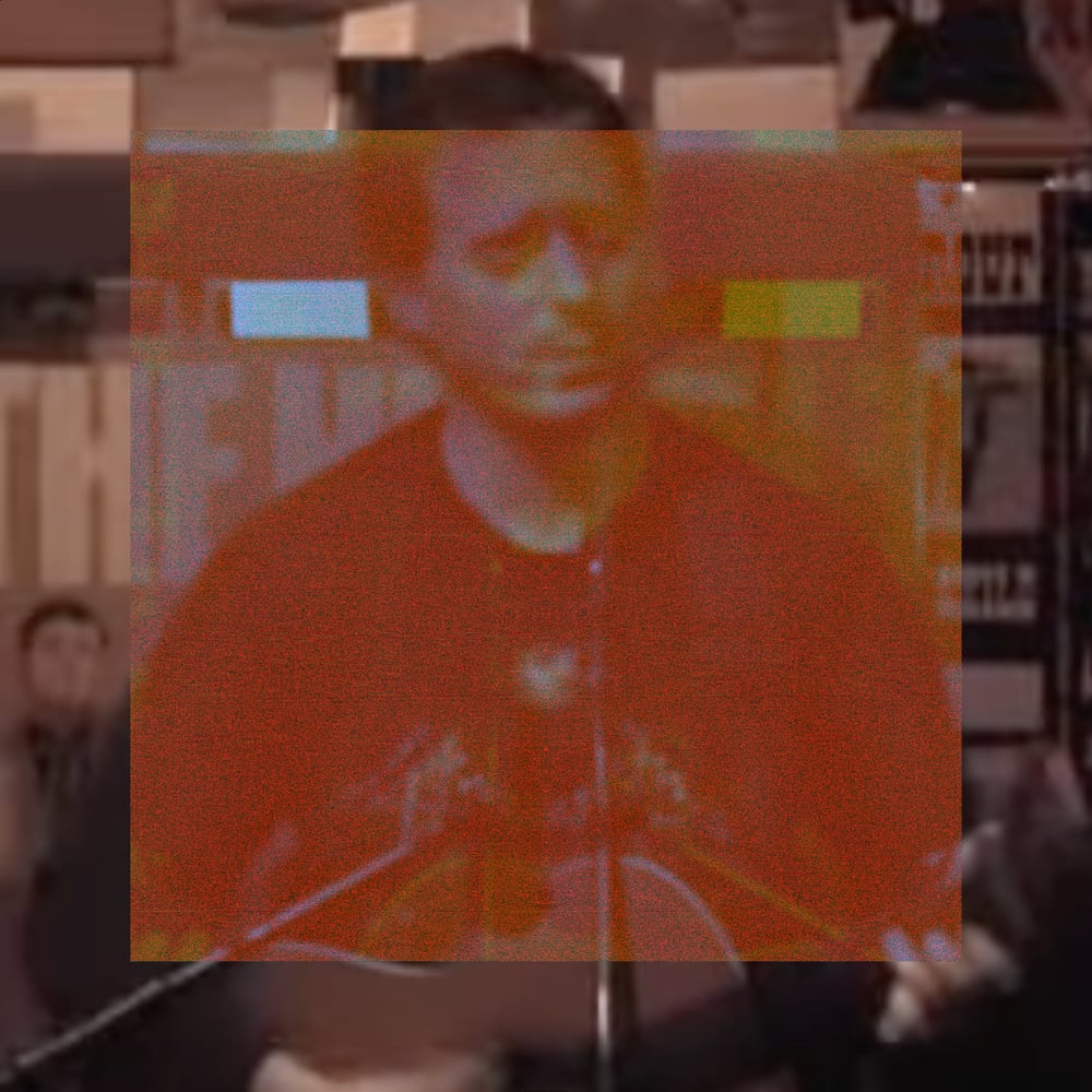A medium shot of Jason Molina seated at a microphone, head bowed downward, in a record store with shelves of records and posters visible in the background. A large orange-red rectangle covers the central two-thirds of the image in warm distortion, with two small isolated bright blocks, one pale blue and one yellow, like corrupted color artifacts against the orange wash.
